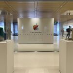 Museum-like Apple exhibit with vintage computers in glass cases, colorful scribbled Apple logo and 50 Years of Thinking Different text on central wall, people walking in bright modern hallway