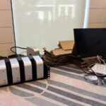 Desk setup with rack-mounted Mac Minis, small monitor, white keyboard, network switch, cables, cardboard trays, and stacked Mac mini boxes against a bright window background