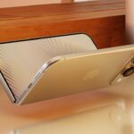 Gold smartphone floating horizontally on a glossy desk, screen glowing with subtle rainbow lines, dual rear cameras visible, beside a small round cat-faced nightlight and wooden background