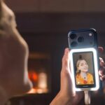 Woman holding a smartphone with a bright rectangular ring light, filming herself in selfie mode; her face appears on the phone screen against a softly blurred indoor background