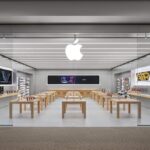 Bright modern Apple retail store with glass front, wooden tables displaying iPhones, iPads, laptops and accessories, illuminated product posters on both walls, and large glowing Apple logo overhead