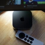 Apple TV box and Siri Remote on a wooden table beneath an LG television displaying a streaming menu with shows like Causeway and Ted Lasso