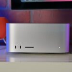 Silver Mac Studio desktop computer on a desk below a monitor, with front ports visible, next to a red smart speaker and other tech accessories against a colorful brick wall background