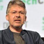 Middleaged man with short gray hair, glasses, and goatee speaking onstage, wearing a dark jacket and headset microphone, with blurred conference text SF 2017 in the background